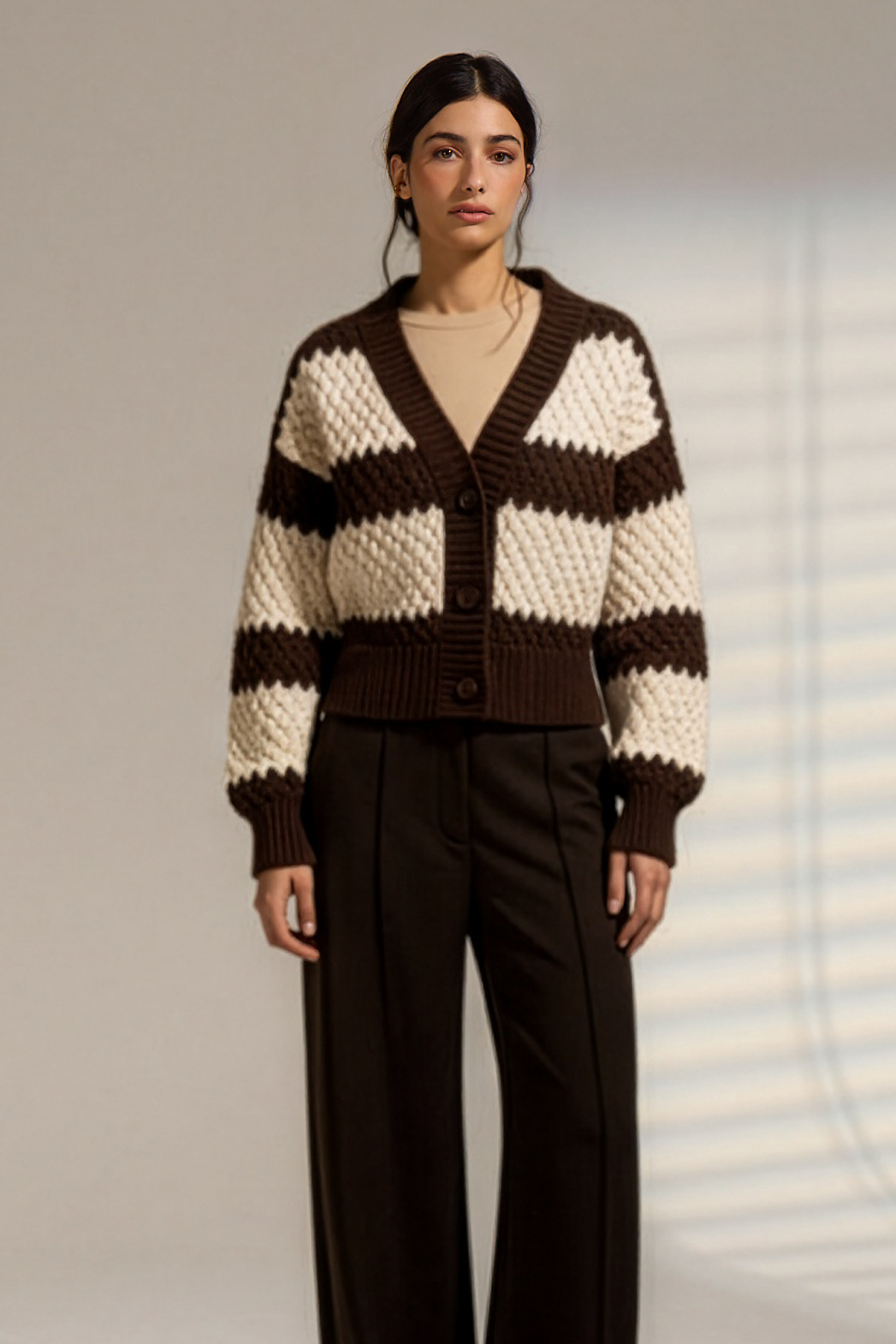 A model stands in a studio wearing a brown and cream striped knit cardigan with a belted waist, paired with dark wide-leg trousers.