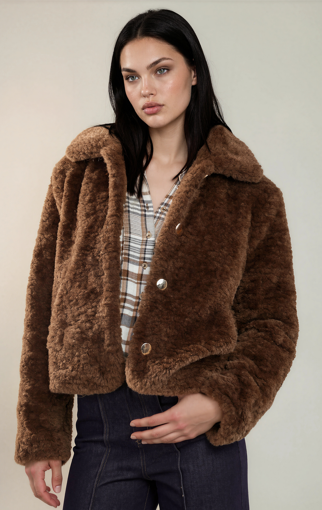 A woman modeling a cropped brown faux-fur jacket with a large collar and silver-tone buttons, worn over a plaid shirt and dark jeans against a plain light beige studio background.