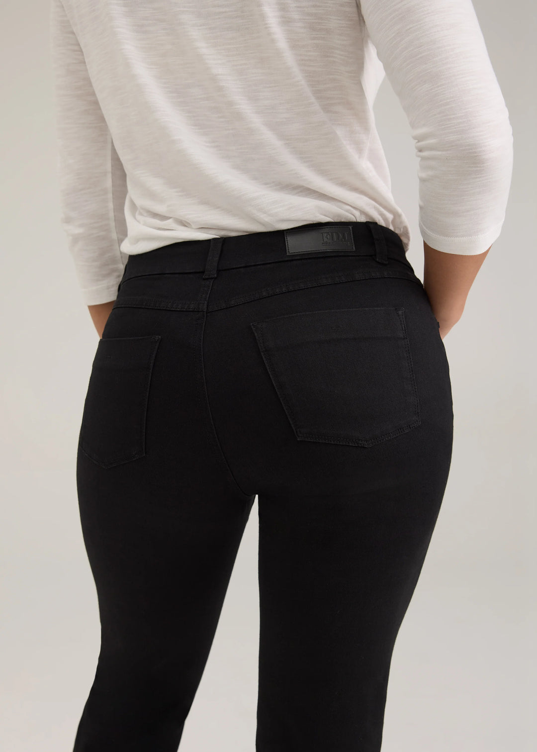 Rear view of a person wearing black high-waisted skinny jeans with a black leather belt and a white long-sleeve top, against a light gray studio background.