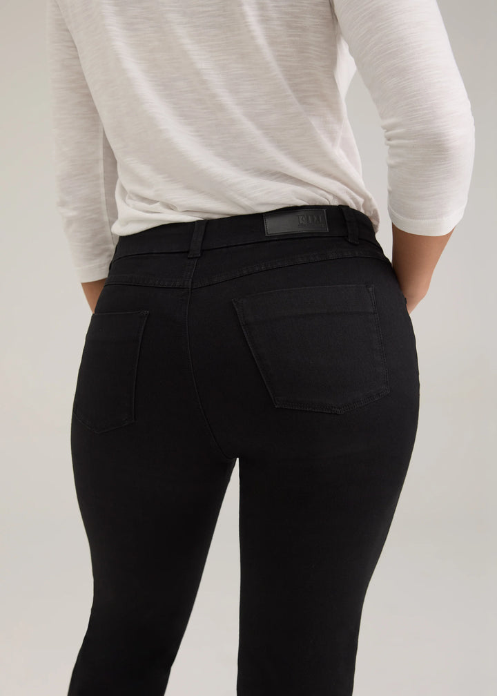 Rear view of a person wearing black high-waisted skinny jeans with a black leather belt and a white long-sleeve top, against a light gray studio background.