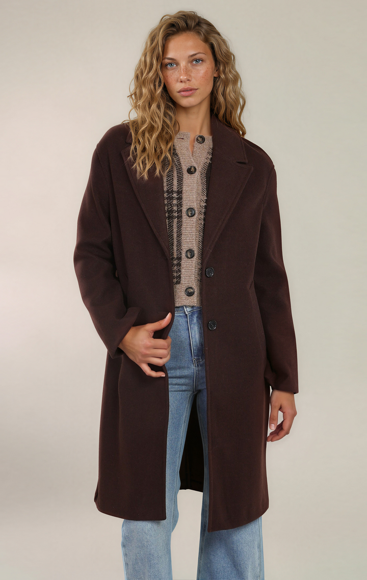 A woman modeling a long, dark brown wool-blend coat over a beige plaid button-front cardigan and light-wash blue jeans, photographed against a plain light beige studio background.