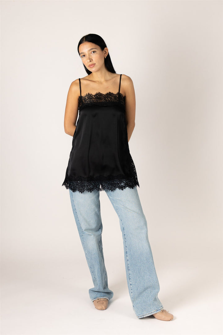 A woman modeling a black satin camisole top with lace trim at the neckline and hem, paired with light-wash wide-leg jeans, against a plain light beige studio background.