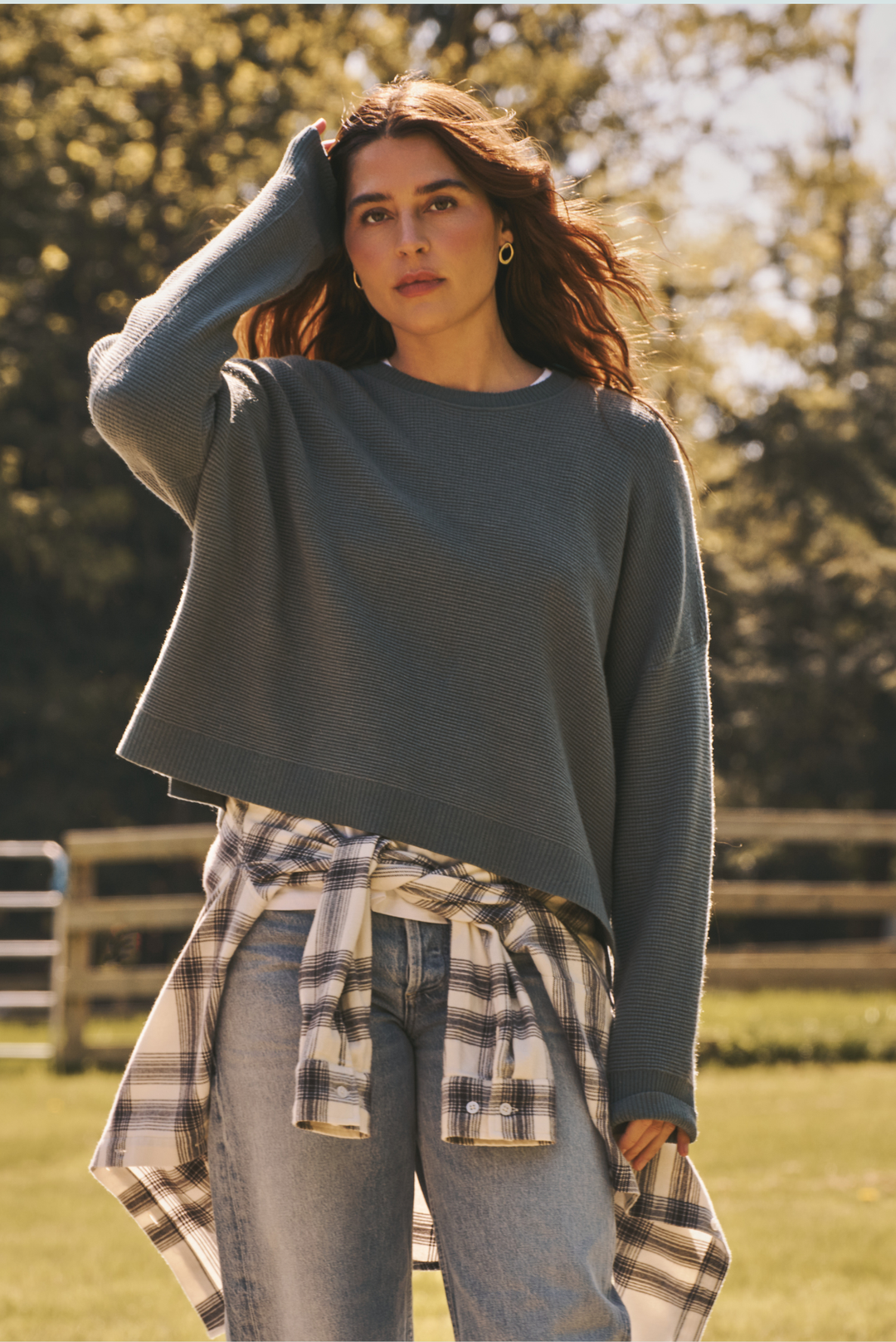 A woman standing outdoors in a grassy field wearing a slate blue cropped long-sleeve sweater, light-wash blue jeans, and a plaid shirt tied around her waist.