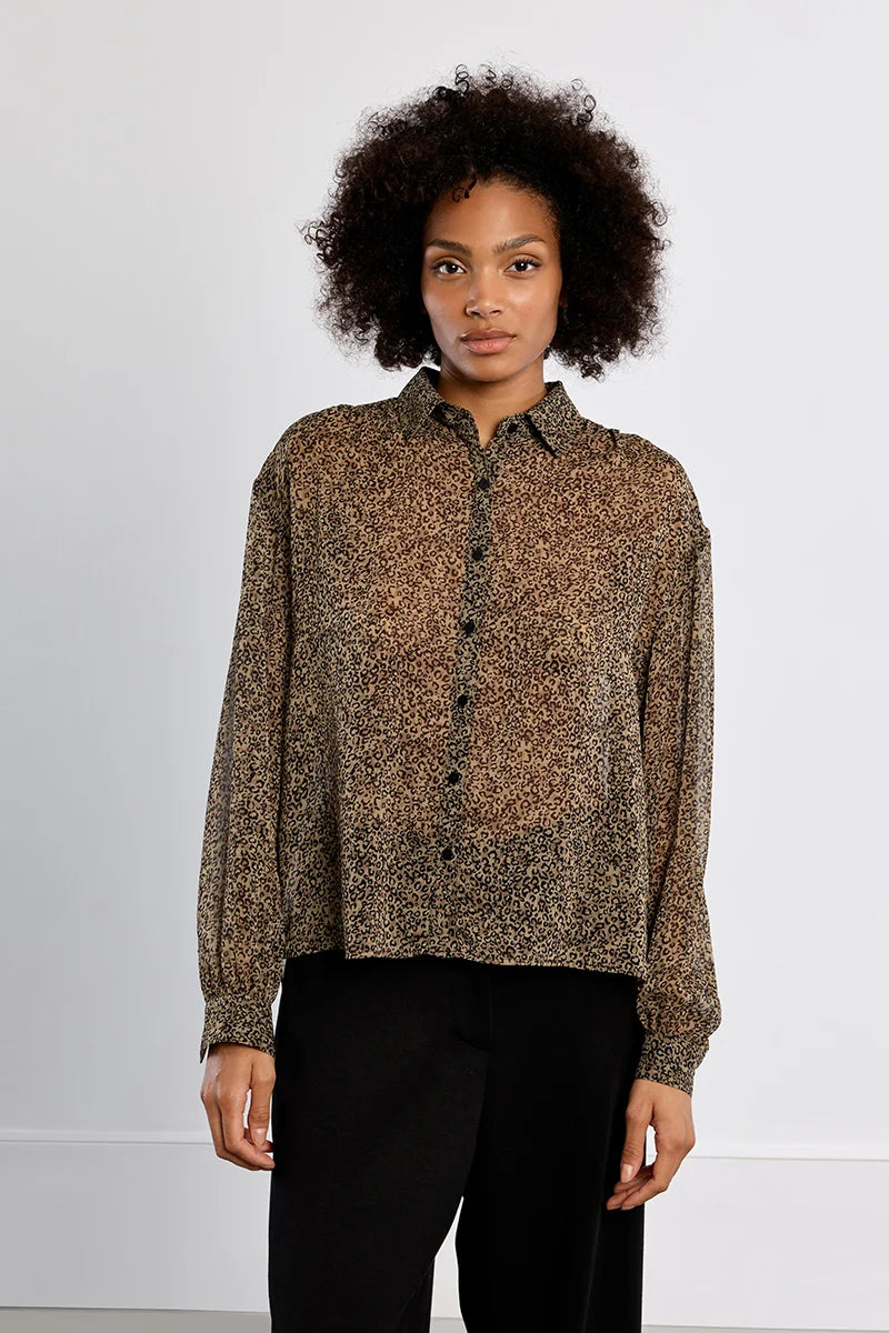 A woman with curly dark hair wearing a brown and black leopard-print long-sleeve button-up shirt and black trousers, standing against a plain white studio background.
