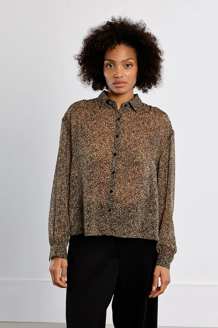 A woman with curly dark hair wearing a brown and black leopard-print long-sleeve button-up shirt and black trousers, standing against a plain white studio background.