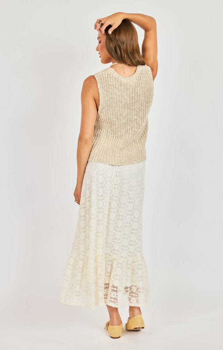 Woman wearing a beige sleeveless top and white lace skirt on a plain background