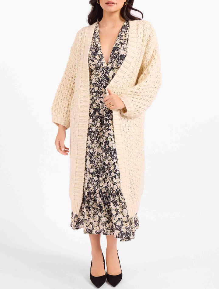 A woman models a long cream-colored chunky knit cardigan over a black and beige floral midi dress, paired with black pointed-toe heels, against a plain white studio background.