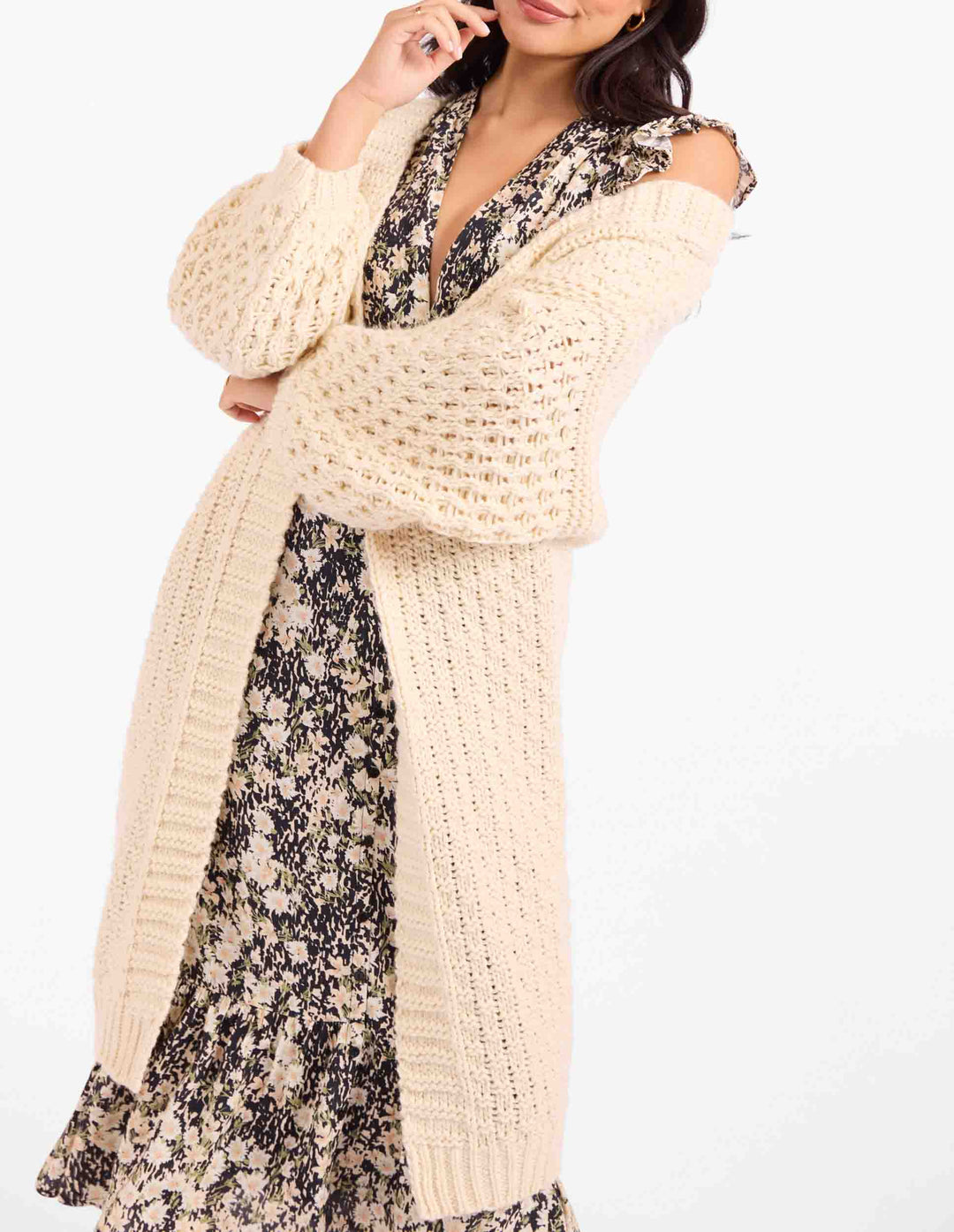 A woman wearing a chunky cream open-knit cardigan over a black and beige floral dress, photographed against a plain white studio background.