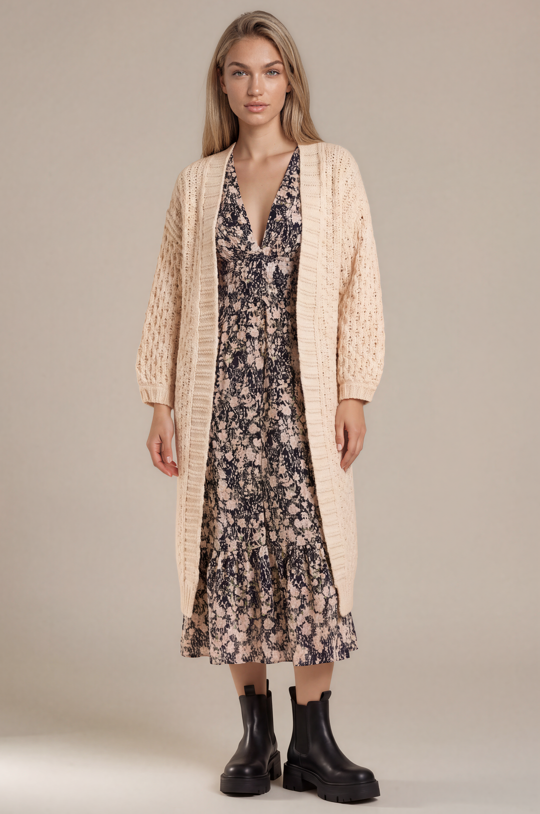 A woman stands against a plain beige studio background wearing a cream cable-knit cardigan over a dark floral midi dress with black ankle boots.