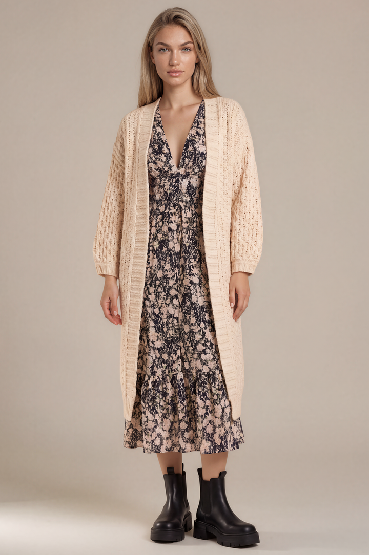 A woman stands against a plain beige studio background wearing a cream cable-knit cardigan over a dark floral midi dress with black ankle boots.