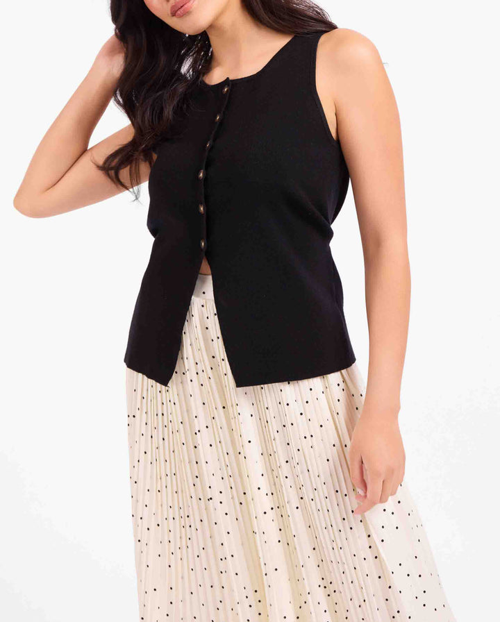 A woman wearing a sleeveless black knit top with decorative buttons down the front and a cream pleated skirt with small black polka dots, photographed against a plain light background.
