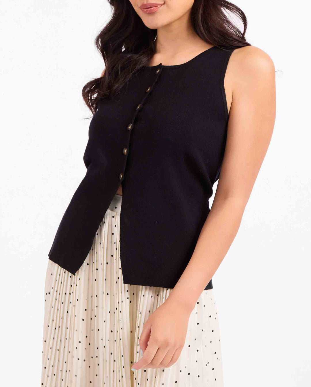 A woman wearing a sleeveless black knit top with a buttoned front and a cream pleated skirt with small black polka dots, photographed against a plain light background.