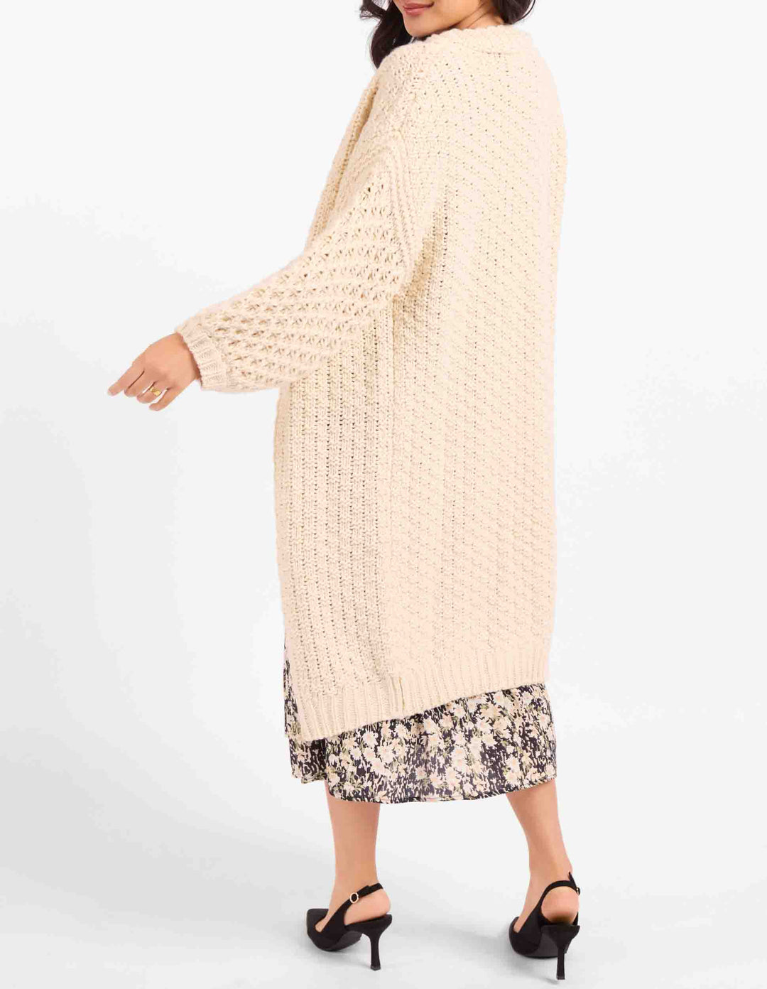 A woman wearing a chunky cream-colored open-front knit cardigan over a floral midi skirt, paired with black high-heeled sandals, shown from a three-quarters back view against a plain white studio background.