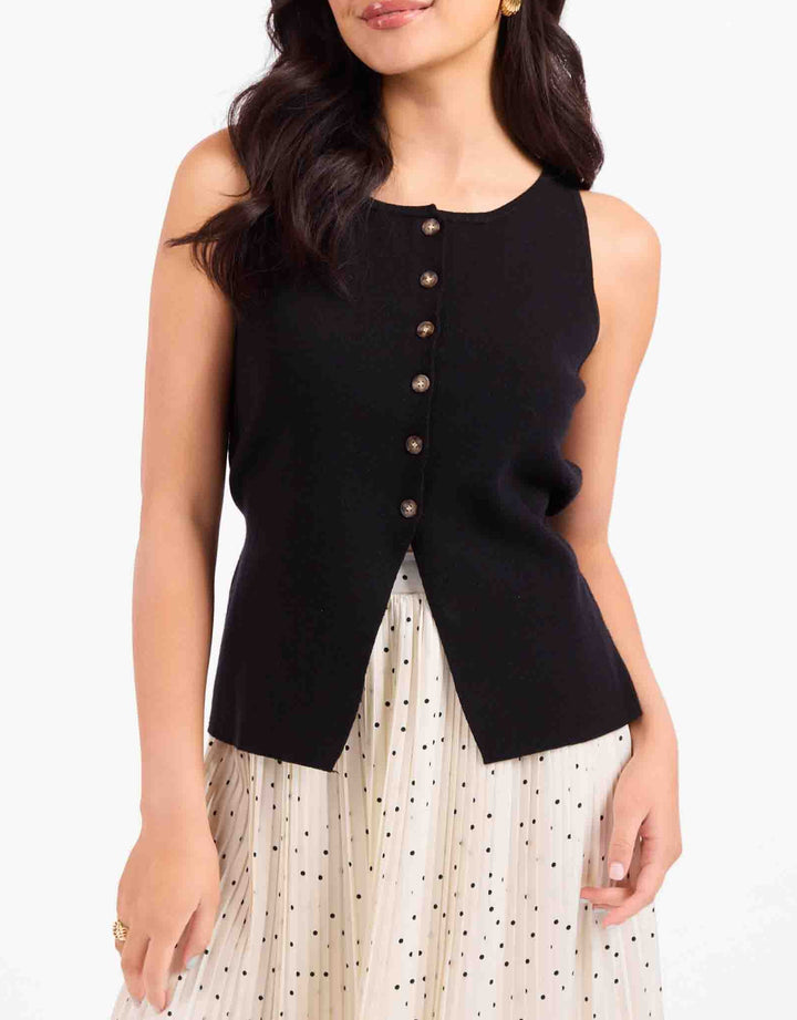 A woman wearing a sleeveless black knit top with a button-front detail and a cream pleated skirt with small black polka dots, photographed against a plain light background.