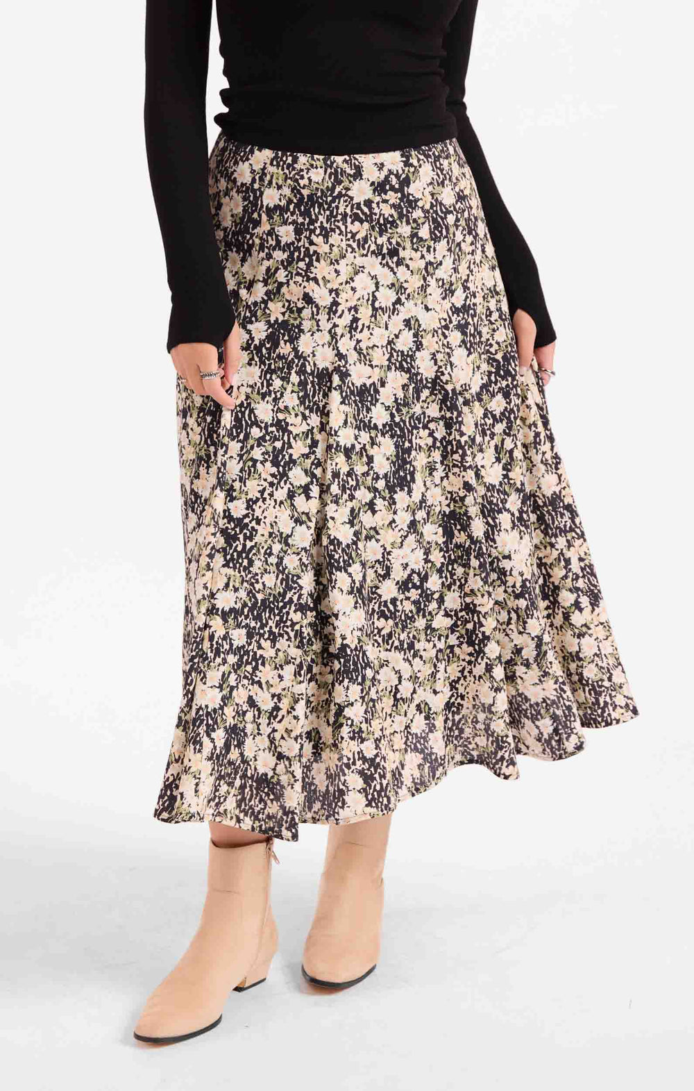 A woman wearing a black long-sleeve top and a knee-length floral midi skirt with beige ankle boots, shown against a plain light gray studio background.