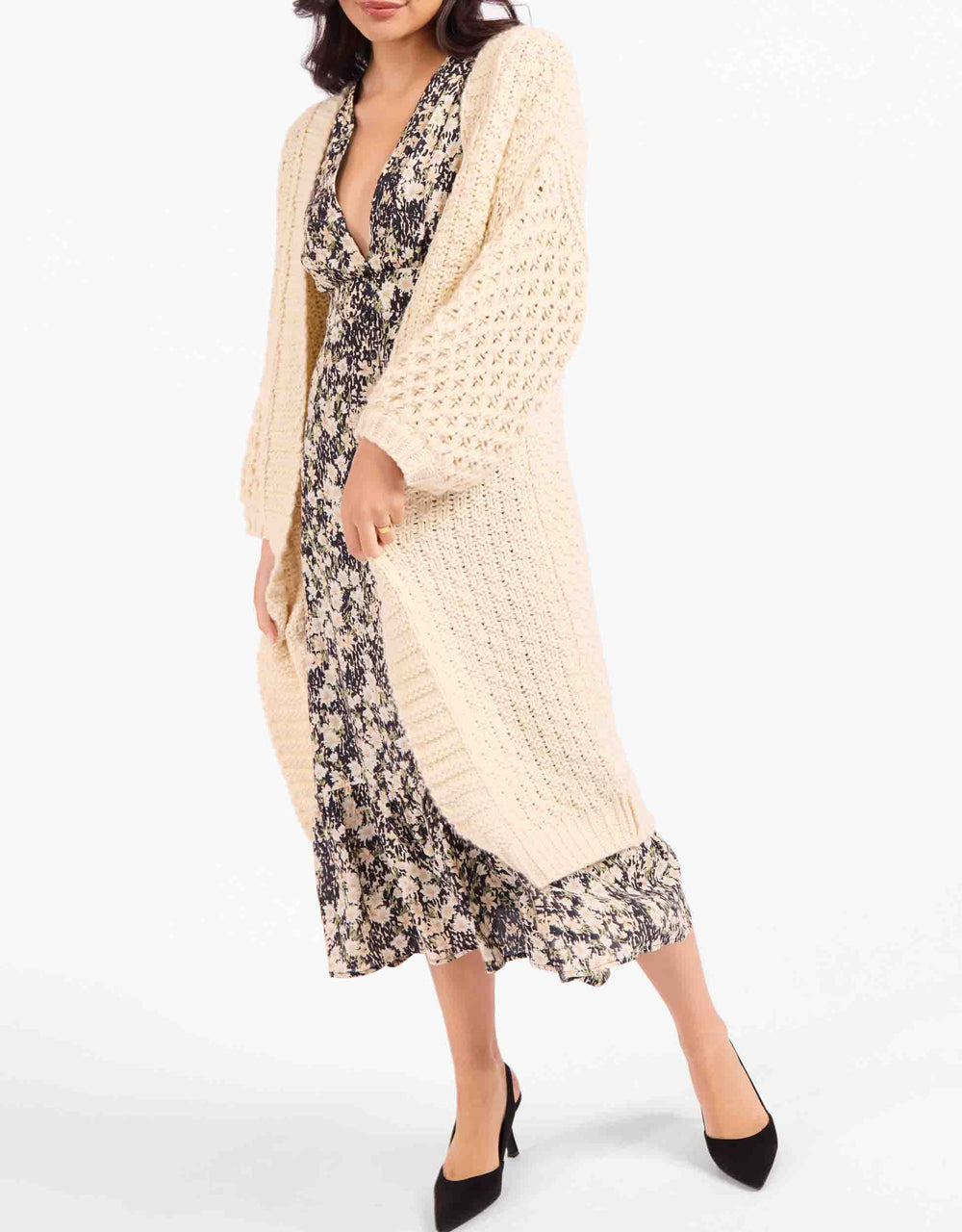 A woman models a long cream open-front cardigan over a dark floral midi dress with black pointed-toe heels, photographed against a clean white studio background.