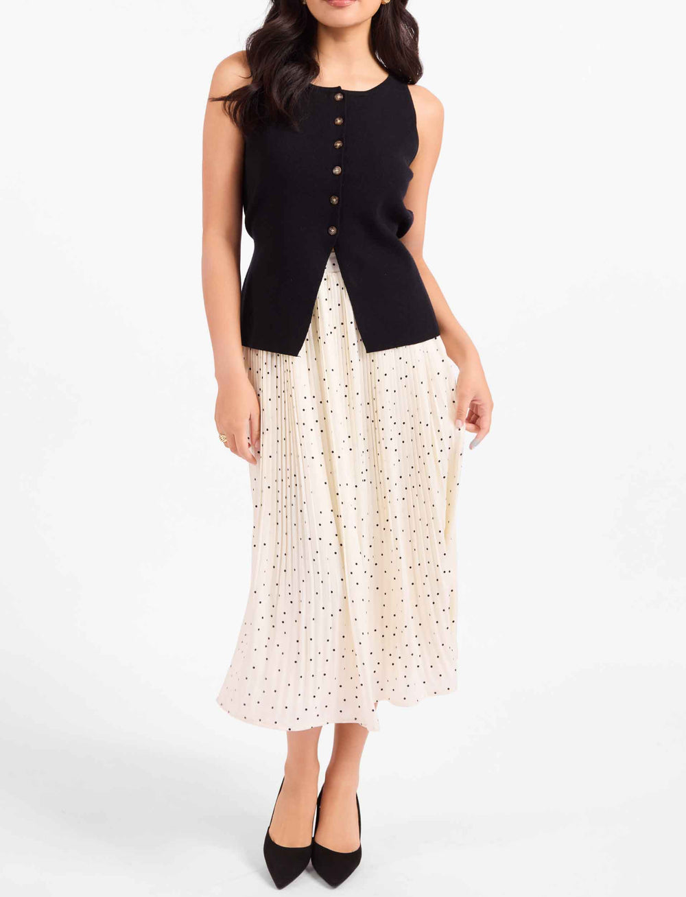 A woman modeling a sleeveless black knit vest with gold-tone buttons over a cream eyelet midi skirt with small black polka dots, paired with black pointed-toe heels, against a plain white studio background.