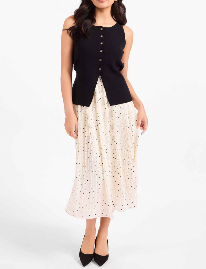 A woman modeling a sleeveless black knit vest with gold-tone buttons over a cream eyelet midi skirt with small black polka dots, paired with black pointed-toe heels, against a plain white studio background.