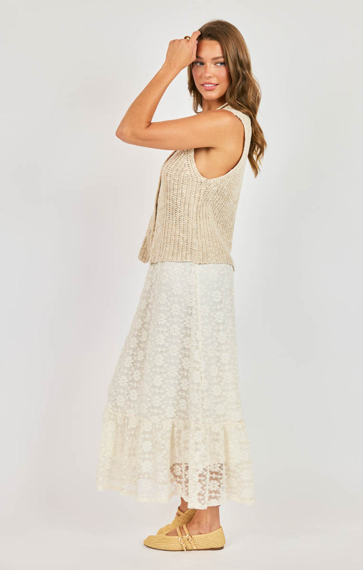 Woman wearing a beige sleeveless top and white lace skirt on a plain background