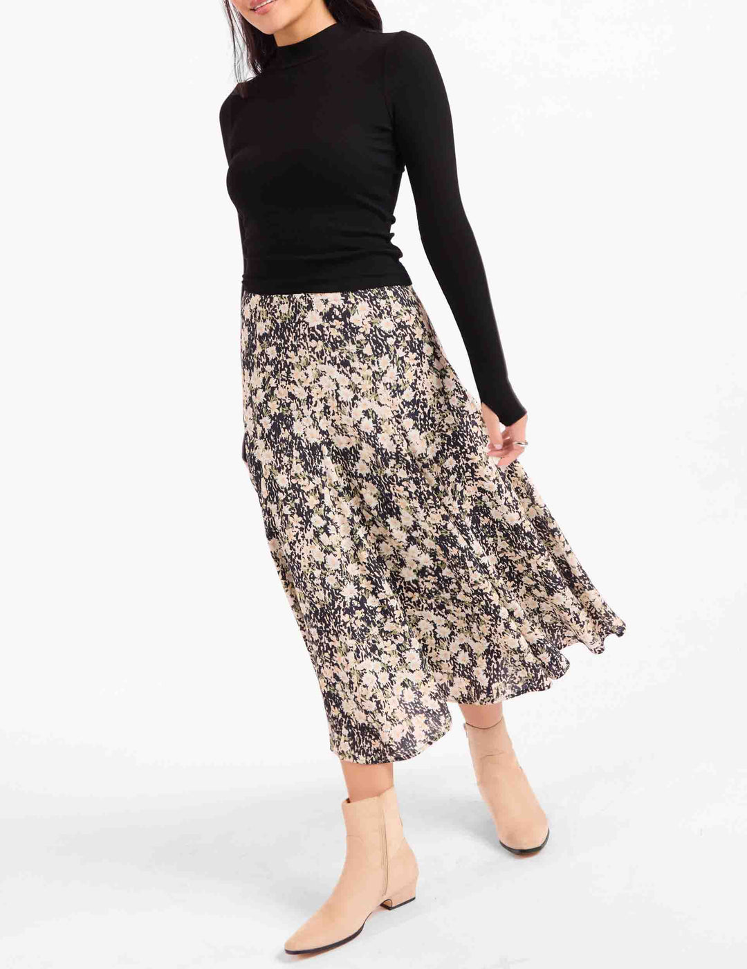 A woman wearing a black long-sleeve mock-neck top and a knee-length floral midi skirt with beige ankle boots, photographed against a plain white studio background.
