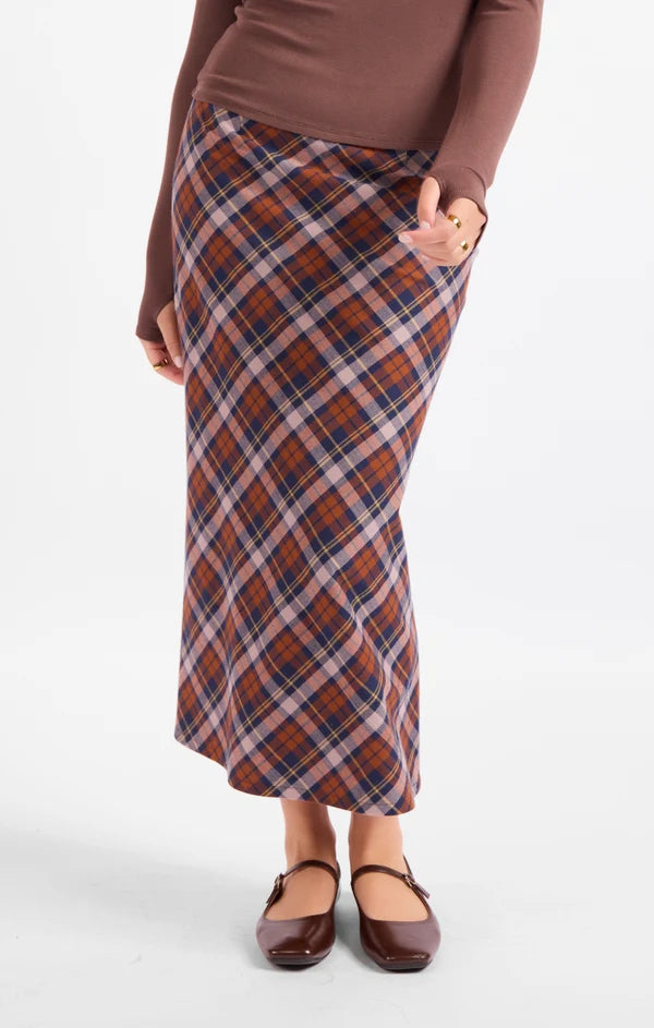 A woman wearing a brown long-sleeve top and a rust-orange plaid midi skirt with navy and white stripes, paired with brown Mary Jane flats, against a plain white studio background.