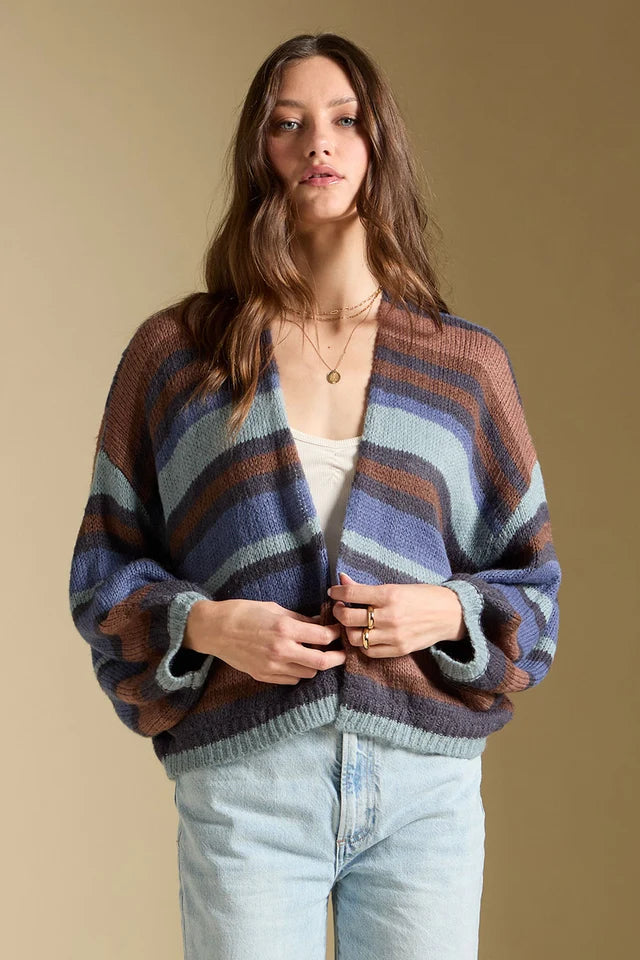 A woman wearing a chunky knit cardigan with horizontal stripes in brown, blue, and gray, styled with light-wash jeans against a solid beige background.