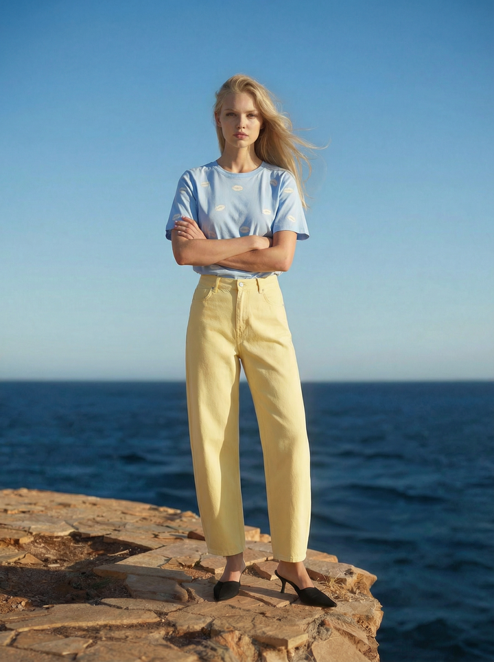 Woman standing on a rocky outcrop by the ocean wearing a light blue t-shirt and yellow pants.