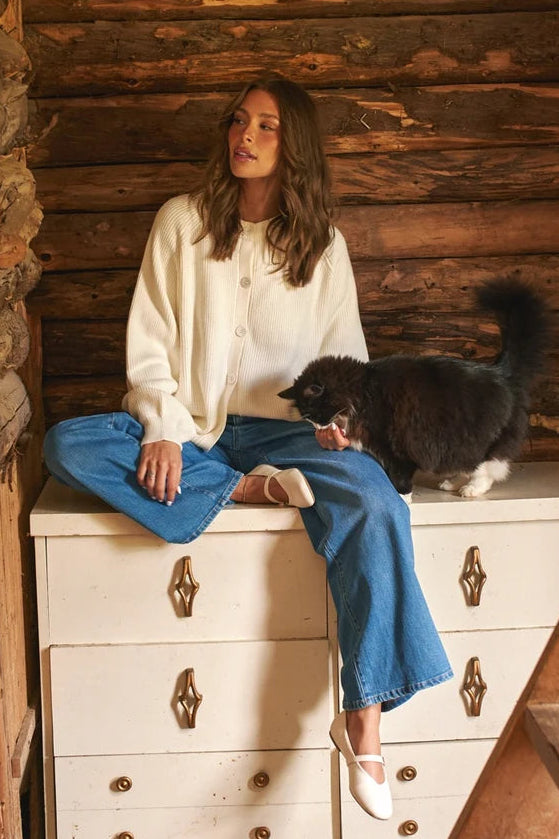 A woman sits cross-legged on a white wooden chest in a rustic log cabin setting, wearing a cream cardigan, blue wide-leg jeans, and white pointed-toe flats. A black and white cat is standing on the chest next to her.