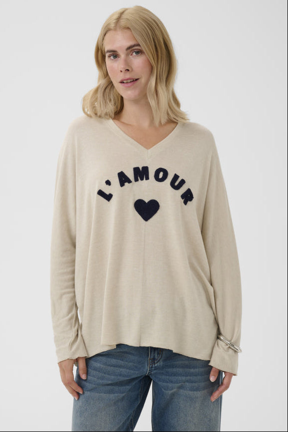 A woman wearing a beige long-sleeve V-neck top with 'L'AMOUR' and a heart printed in navy on the front, paired with blue jeans, against a light gray studio background.