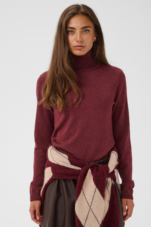 A woman modeling a heathered burgundy turtleneck sweater, styled with a matching burgundy and cream argyle knit wrap tied at the waist, against a light gray studio background.