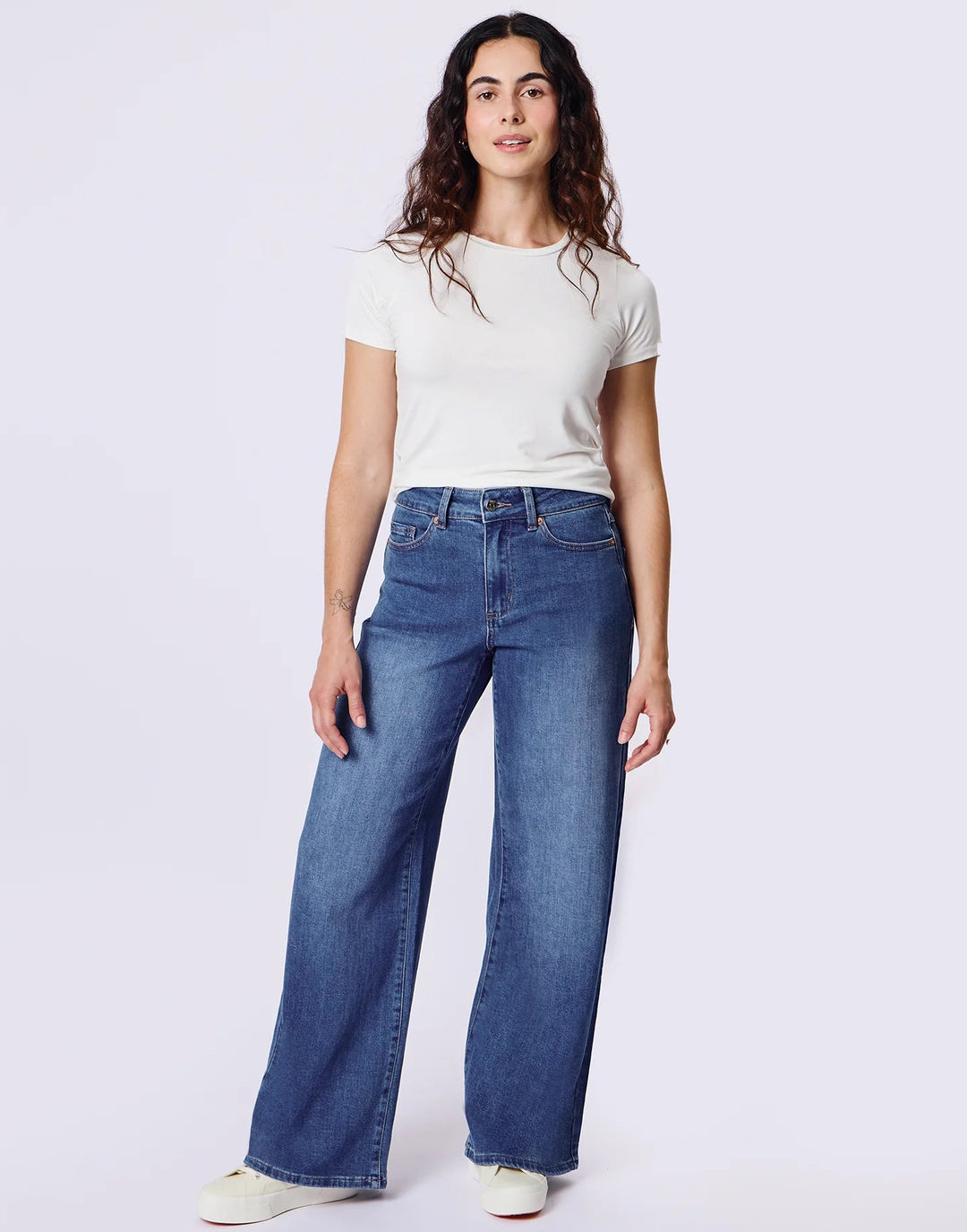 A woman modeling a pair of high-waisted, wide-leg blue jeans with a white t-shirt and white sneakers, standing against a light gray studio background.