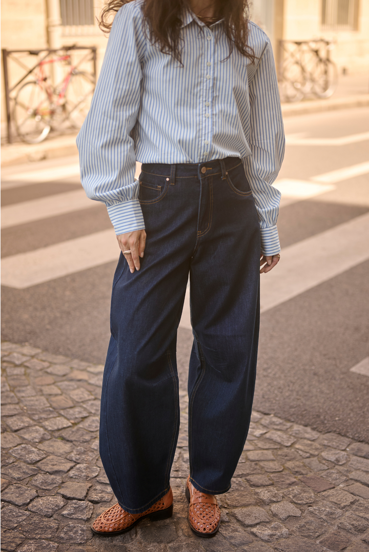 A person standing outdoors on a cobblestone street wearing a light blue and white vertically striped button-up shirt, dark wide-leg denim jeans, and brown woven leather shoes.