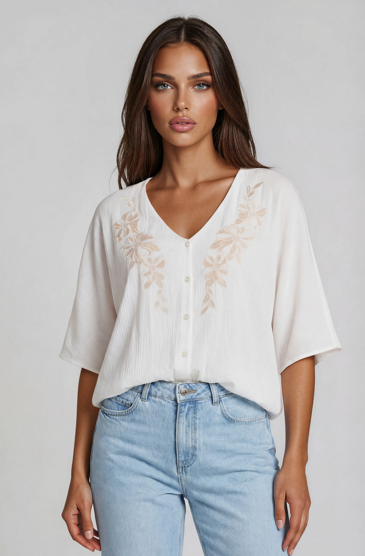 Woman wearing a white blouse with floral embroidery and blue jeans on a plain background