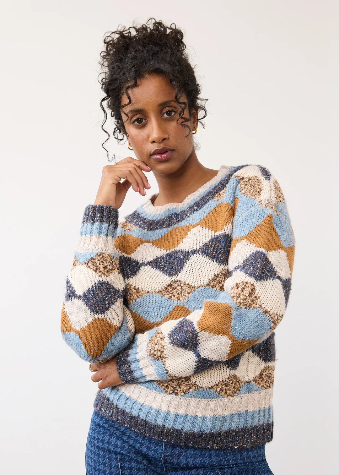 A woman wearing a chunky knit sweater with a geometric diamond pattern in light blue, navy, tan, and cream, paired with blue patterned pants, against a plain light gray studio background.
