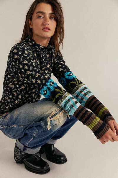 Person wearing a patterned sweater with colorful sleeves and jeans, sitting on the ground.