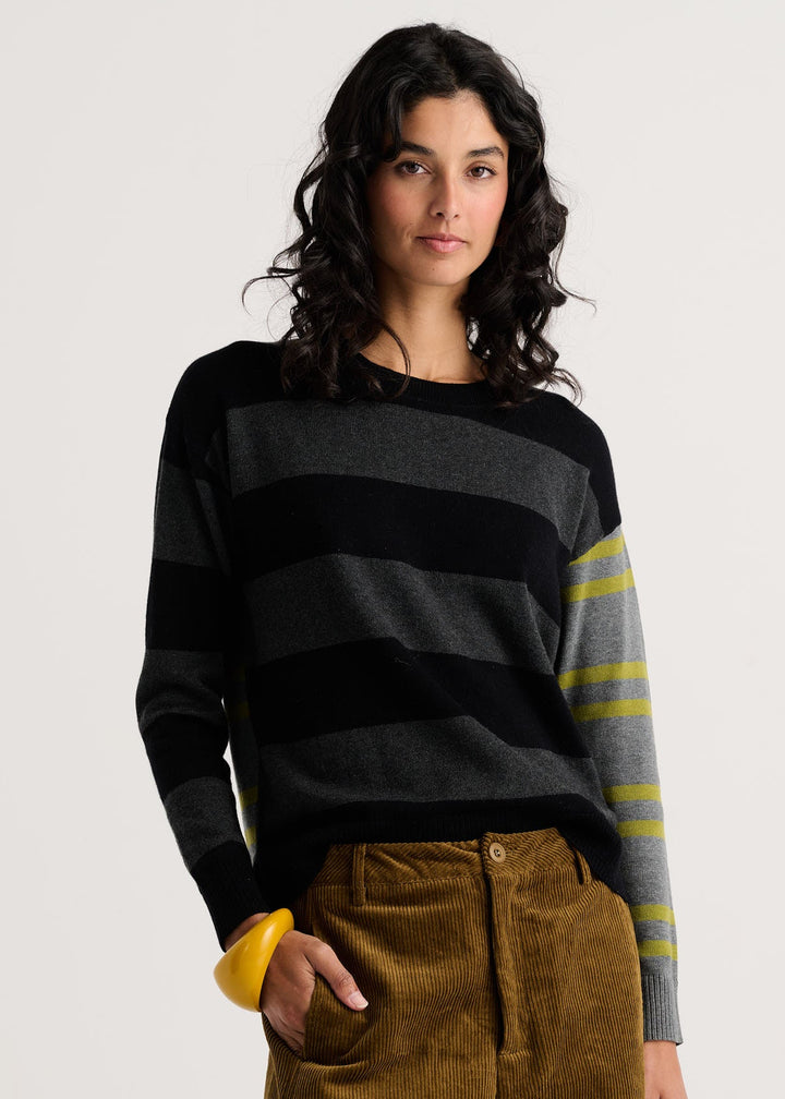 A woman modeling a striped knit sweater with horizontal black and gray stripes and yellow-green stripes on the sleeves, paired with brown corduroy pants, against a light gray studio background.