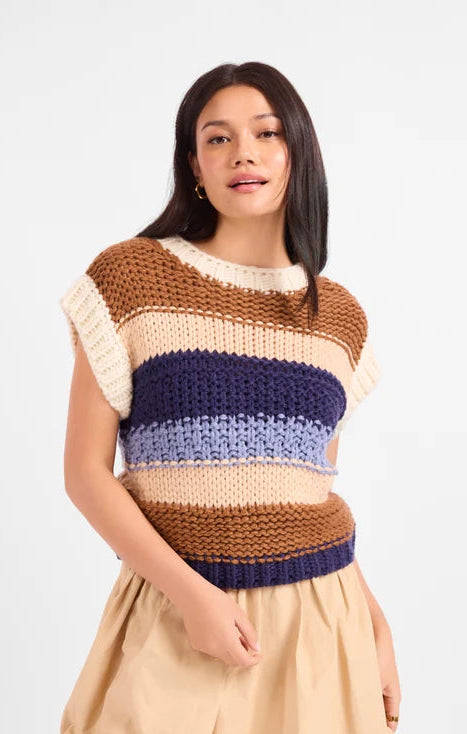 A woman modeling a chunky knit sleeveless top with horizontal stripes in brown, cream, navy, and light blue, paired with beige shorts, against a light gray studio background.