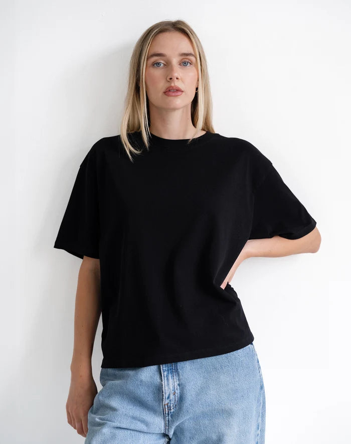 A woman wearing an oversized black short-sleeve t-shirt and light blue jeans, standing against a plain white wall.