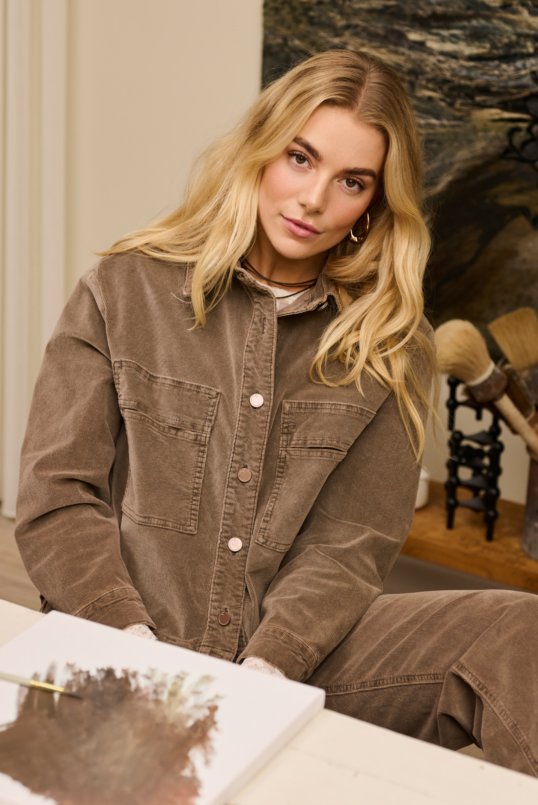 A blonde woman wearing a brown corduroy button-up shirt and matching pants, sitting in a warm, artistic interior setting.
