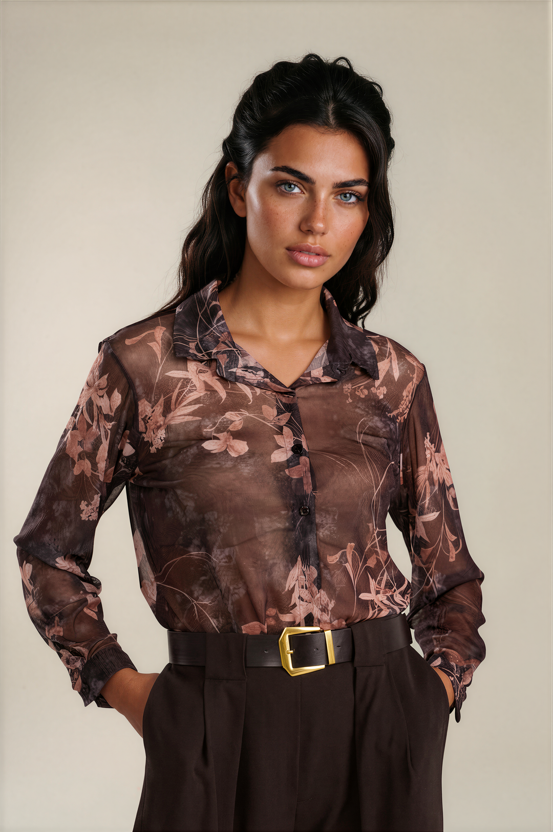 A woman modeling a sheer long-sleeve button-up blouse with a brown and dusty pink floral pattern, tucked into dark brown trousers with a black belt featuring a gold buckle.
