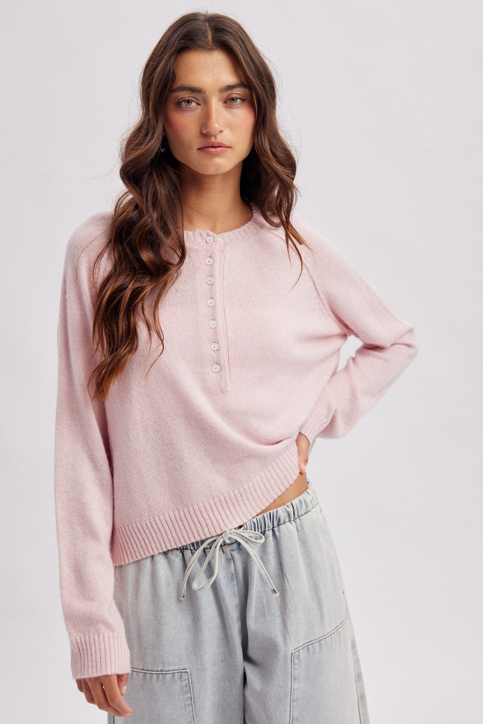 A woman modeling a light pink cropped knit sweater with a buttoned henley-style neckline, paired with light gray drawstring pants, against a plain light gray studio background.