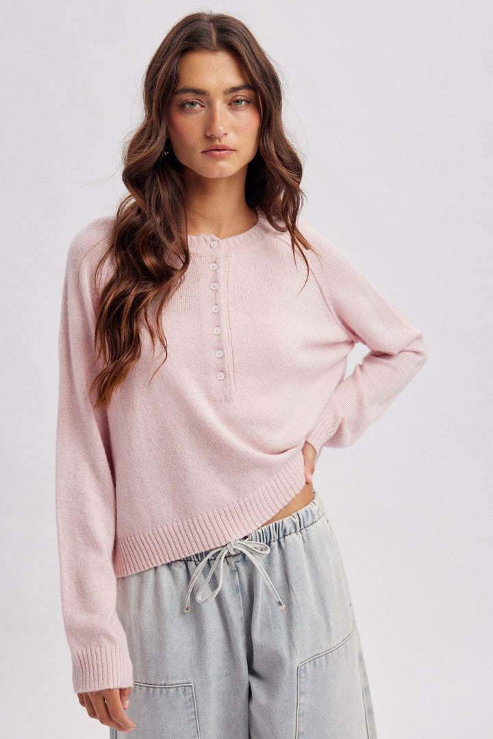 A woman modeling a light pink cropped knit sweater with a buttoned henley-style neckline, paired with light gray drawstring pants, against a plain light gray studio background.