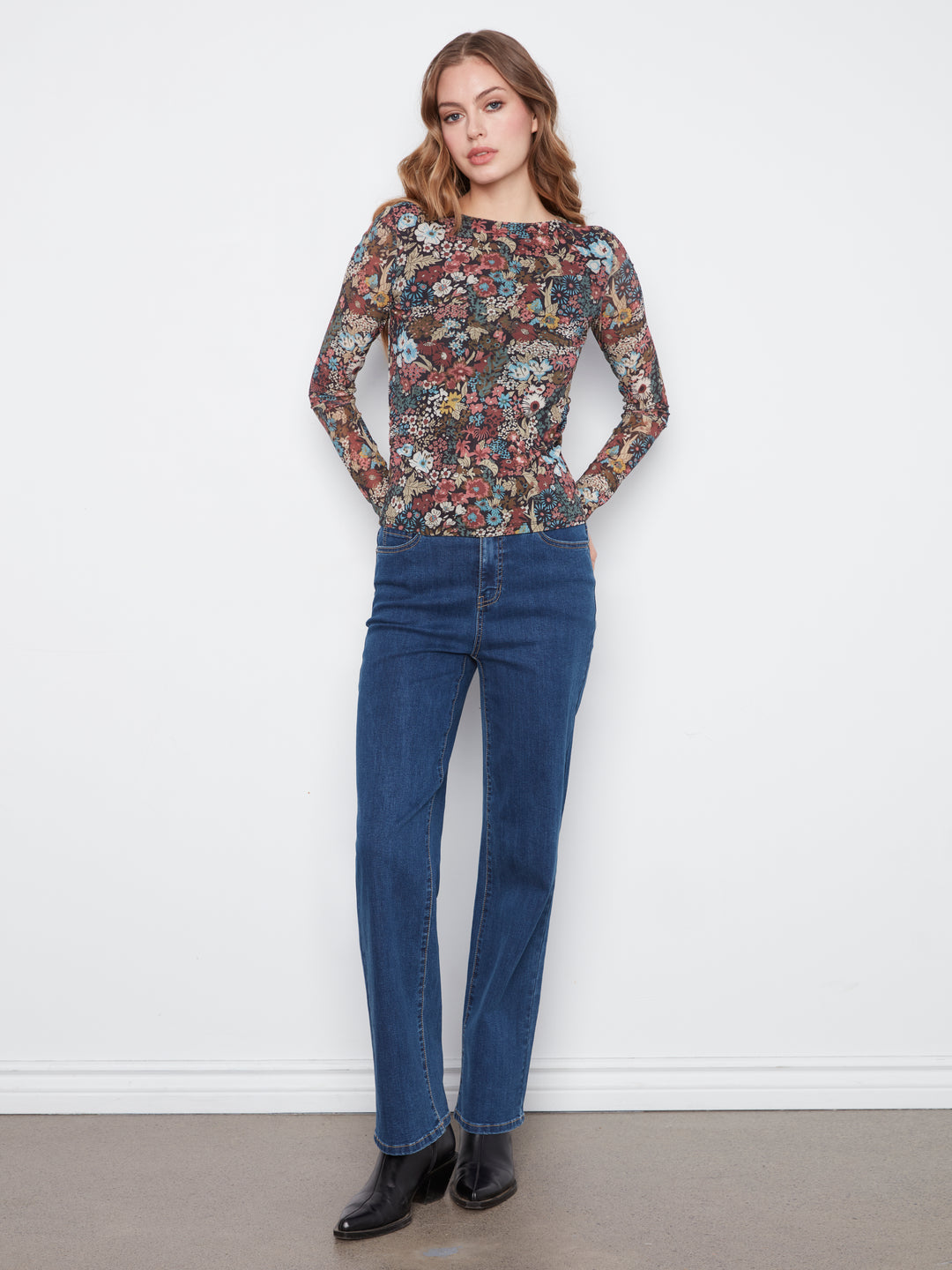 A woman modeling a long-sleeve crewneck top with a dense multicolor floral and abstract print, paired with dark blue jeans and black ankle boots, standing against a plain white wall.