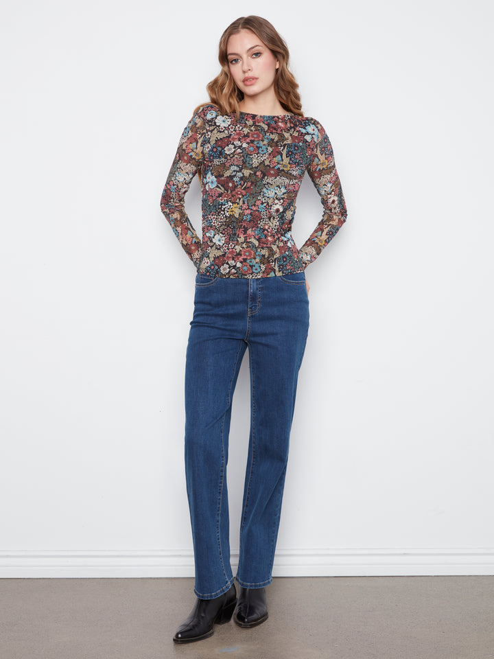 A woman modeling a long-sleeve crewneck top with a dense multicolor floral and abstract print, paired with dark blue jeans and black ankle boots, standing against a plain white wall.