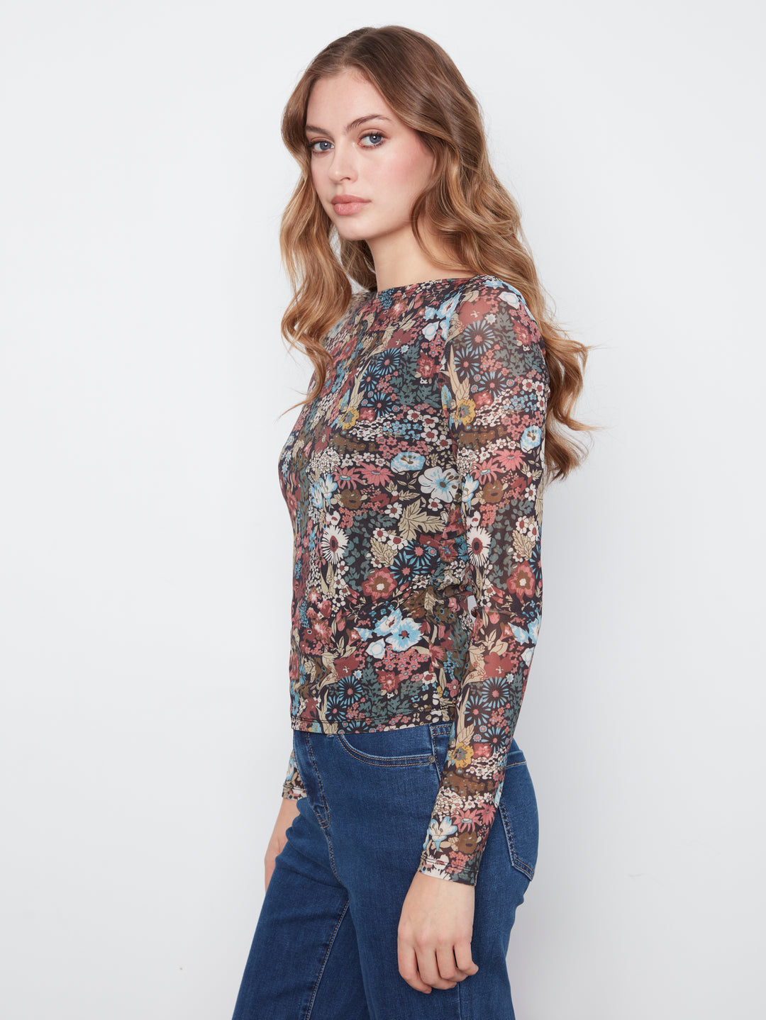 A woman wearing a long-sleeve crewneck top with an intricate multicolor floral and paisley print, paired with blue jeans, photographed against a plain light gray background.