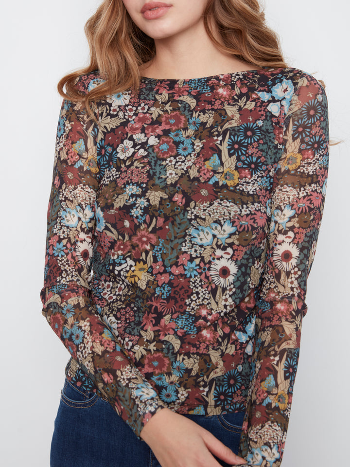A woman wearing a long-sleeve boat-neck top with an intricate multicolor floral and bird print, paired with dark blue jeans, shown against a plain light gray background.
