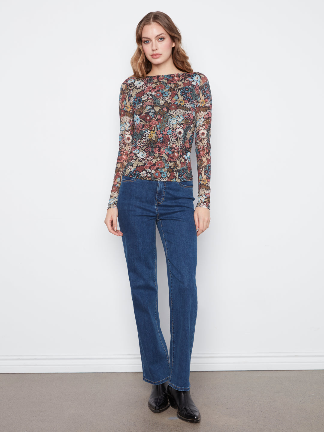 A woman modeling a long-sleeve boat-neck top with a dense multicolor floral and botanical print, paired with dark blue jeans and black boots, standing against a plain white wall.