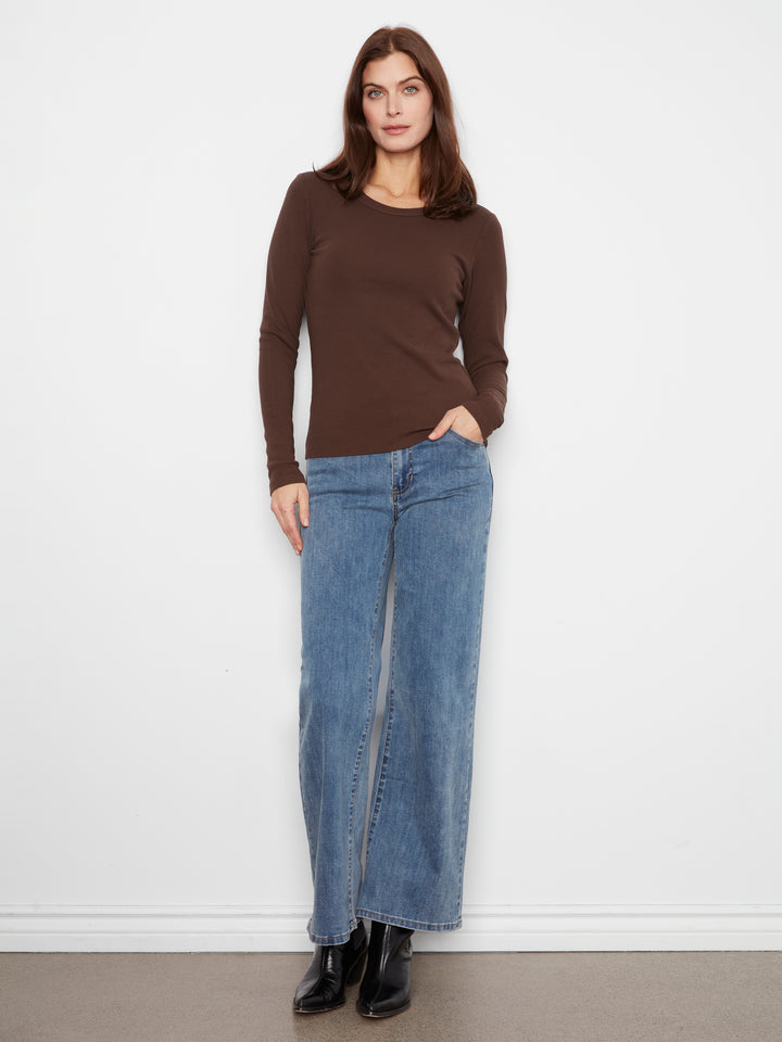 A woman modeling a brown long-sleeve crewneck top and wide-leg blue jeans with front slits, paired with black ankle boots, standing against a plain white wall.