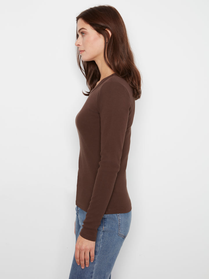 A woman in side profile wearing a fitted, long-sleeve brown V-neck top and blue jeans, standing against a plain white studio background.