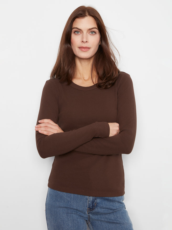 A woman with long brown hair wearing a dark brown long-sleeve crewneck sweater and blue jeans, standing against a plain white studio background with her arms crossed.
